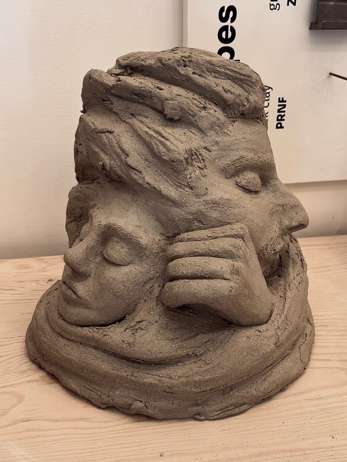 A mounded form in dark grey-brown clay with faces emerging from it: one in profile with closed eyes, a smaller face below, and a hand pressed across the lower face. Coiled ridges of clay wrap the base.