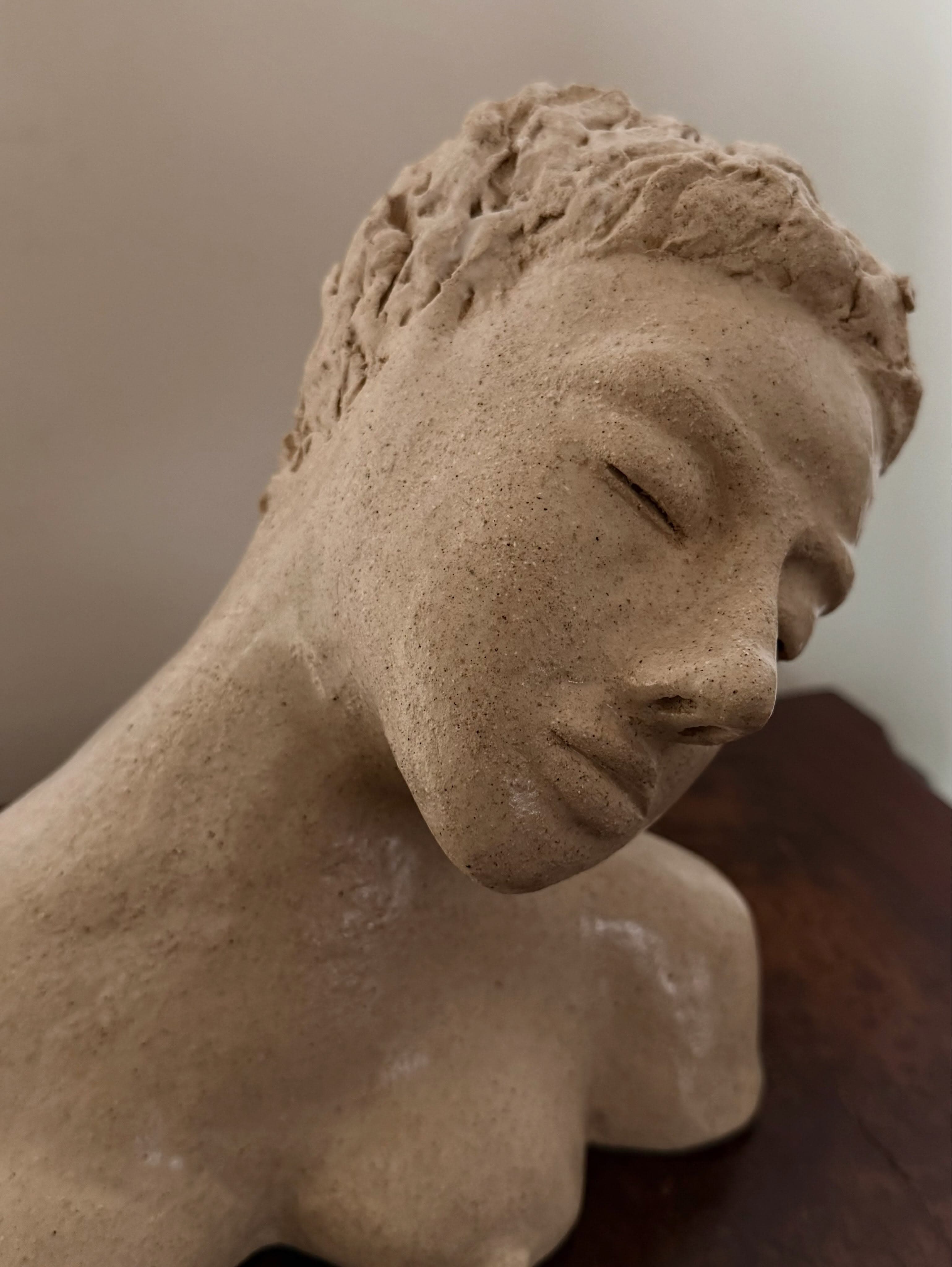 A bust of a woman in buff clay — head, neck and bare shoulders — with the head tilted down and to one side, eyes closed and chin near the shoulder. The hair is short and roughly textured.